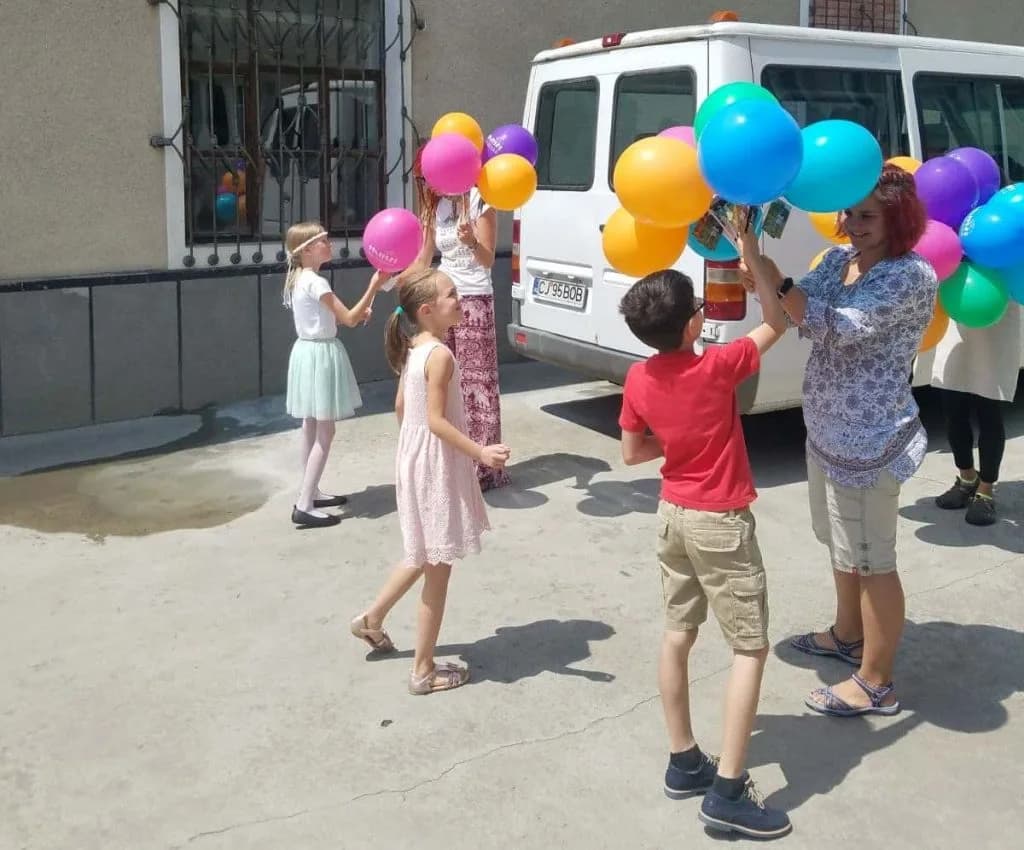 Copii și voluntari țin baloane colorate în curte, lângă microbuz